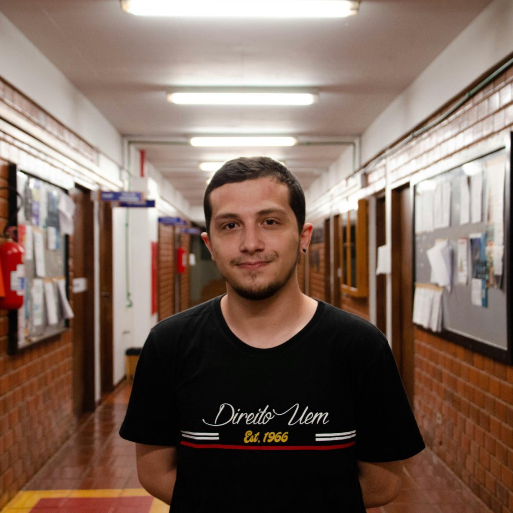 A smiling young man in a black t-shirt stands in a brightly lit academic corridor.