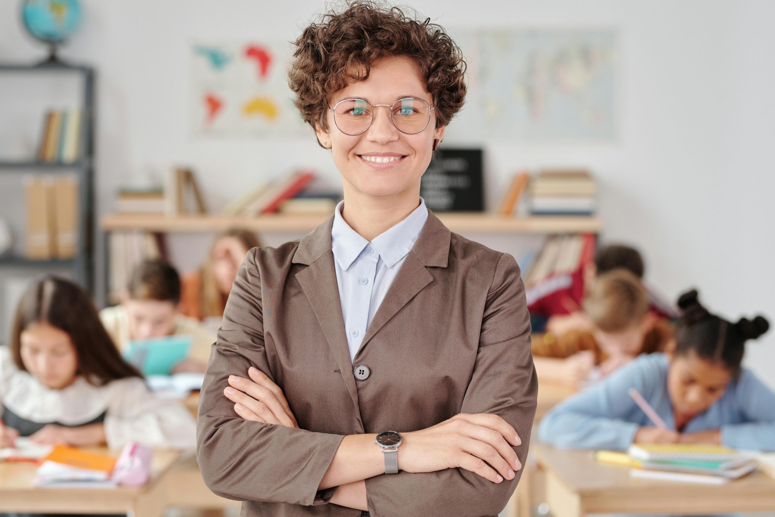 Smiling teacher with glasses standing with arms crossed in a classroom, students learning.