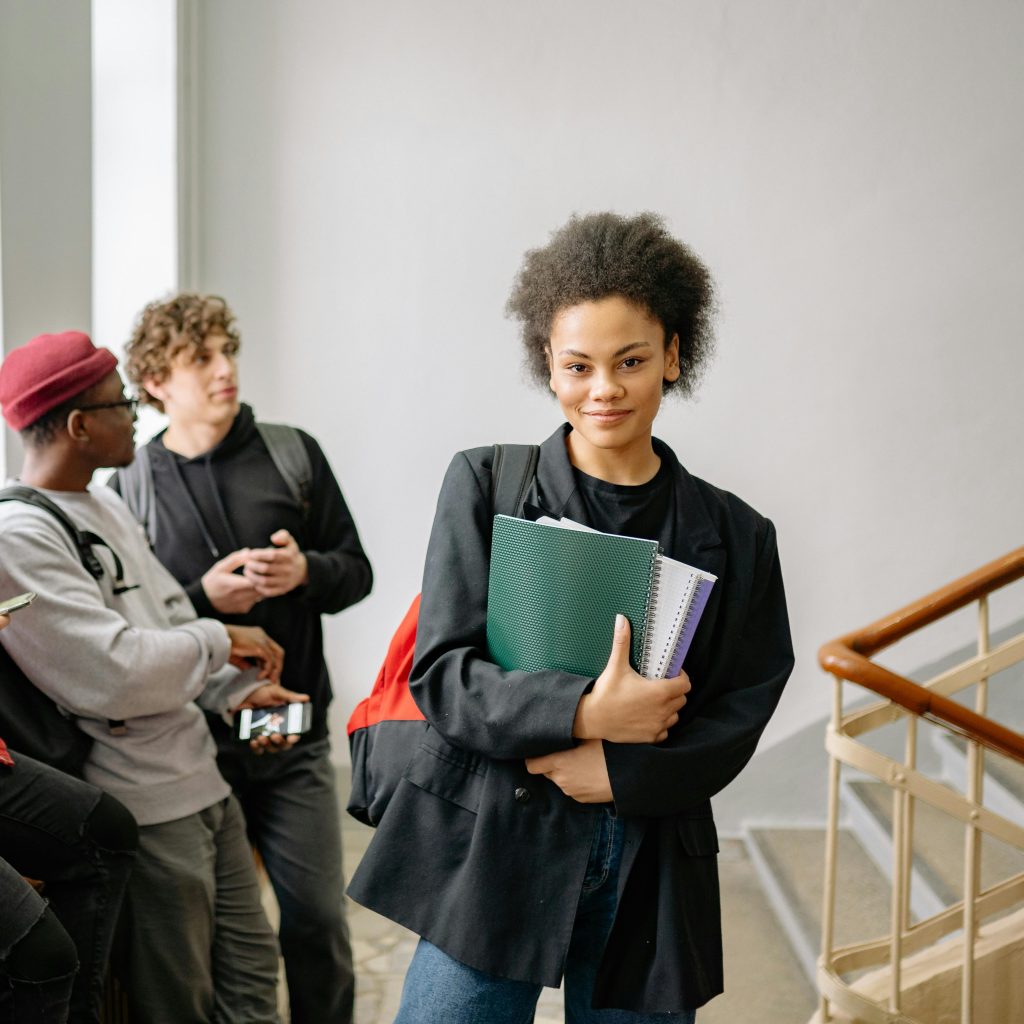 Young woman with afro hair holding notebooks, standing in university hallway, smiling confidently.